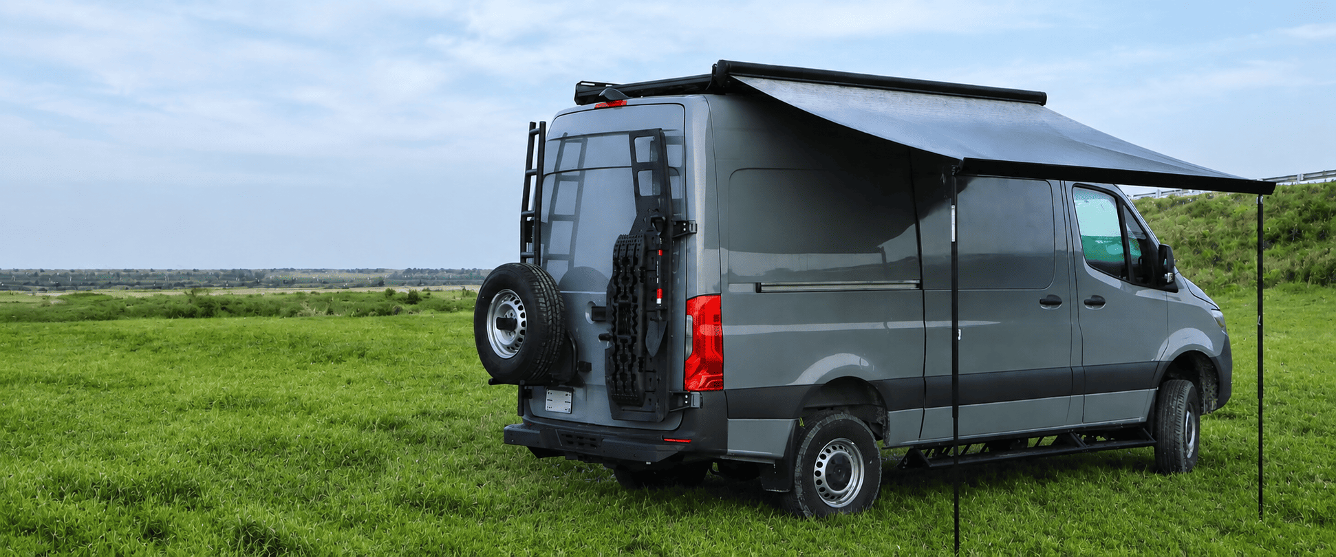 A van with an extended awning set up in a grassy field for outdoor camping or travel.